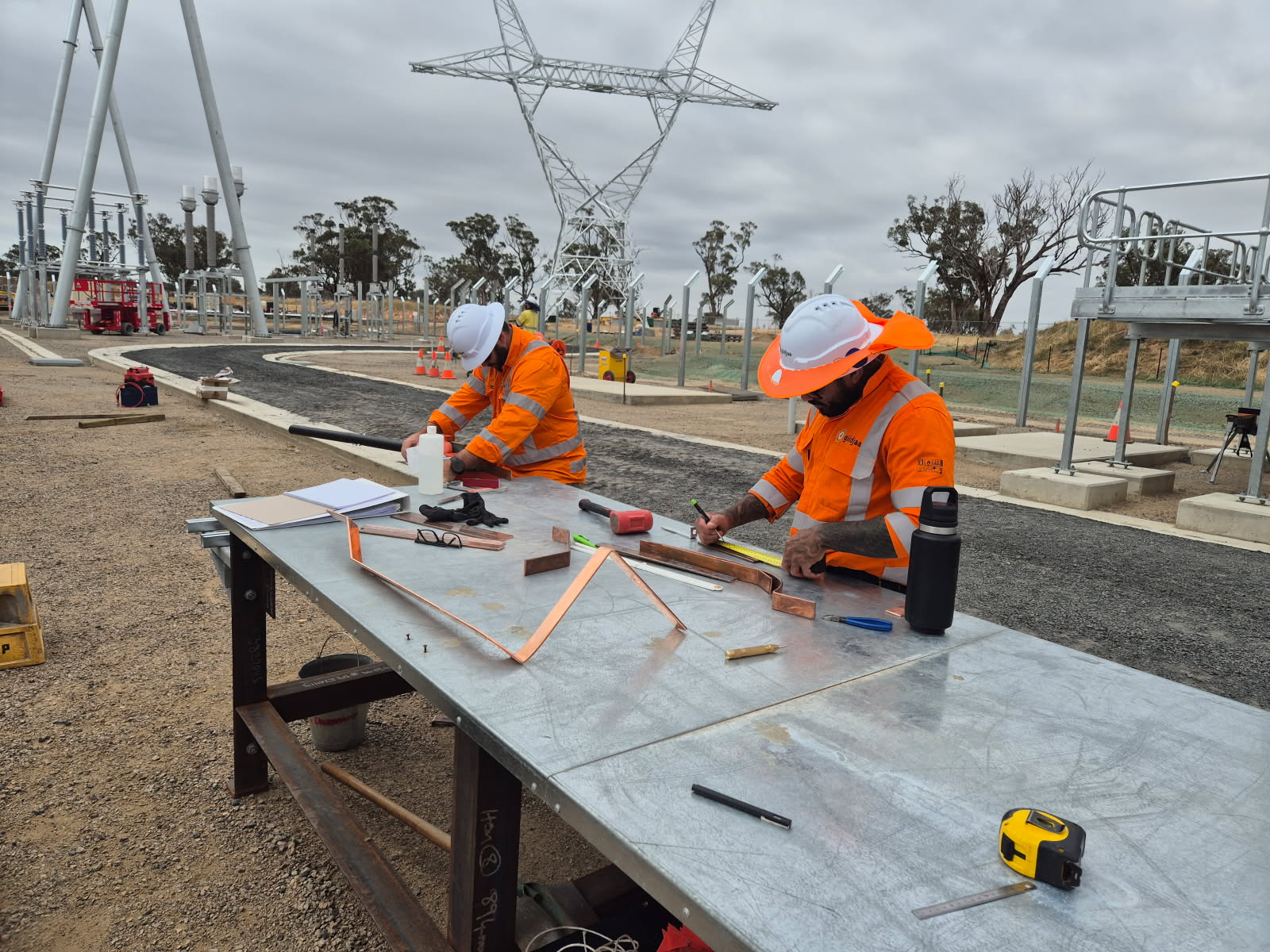 Two Giidjaa electricians in orange hi-vis bending copper earthing bar at a site bench, a transmission tower and the wind farm switchyard behind them